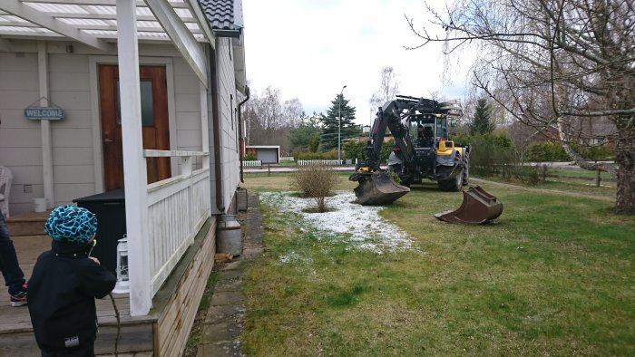 Barn observerar grävmaskin nära hus under byggprojekt på en trädgård med snöfläckar.