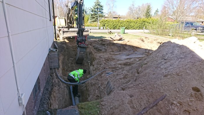 Grävskopa vid husgrund med öppnad grävt för avloppsrenovering, slangar på marken, person i reflekterande jacka arbetar.