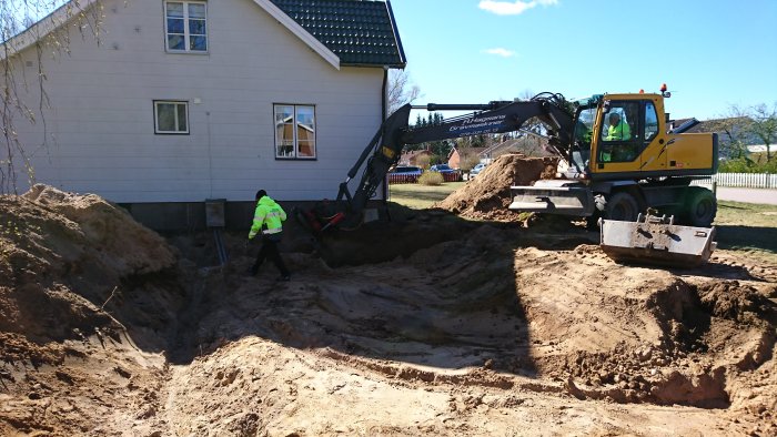 Grävmaskin gräver bredvid en byggnad, person i säkerhetskläder arbetar, stora jordhögar synliga.