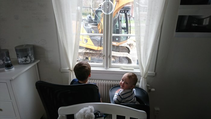 Två barn tittar på en grävmaskin genom ett fönster inifrån ett hus.