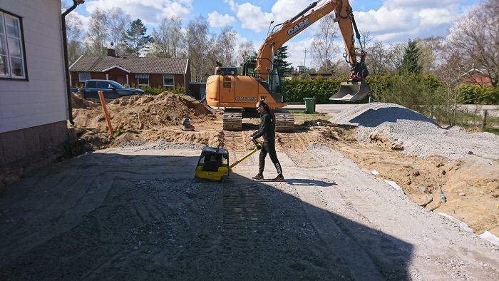 Person använder en handhållen markstamp för att packa makadam medan en grävmaskin och grushögar är i bakgrunden.