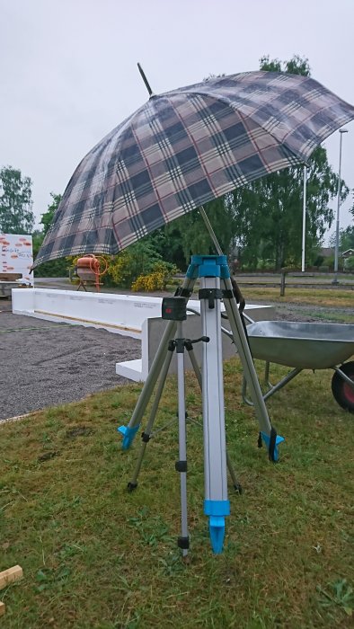 En lasernivå på ett stativ med ett skyddande rutigt paraply ovanför, i en byggarbetsmiljö med blandare och kantelement i bakgrunden.