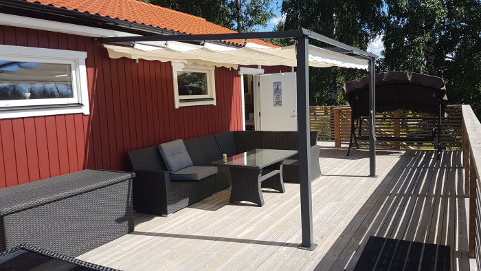 Väggfast pergola monterad på träveranda med möblemang nära röd husfasad.