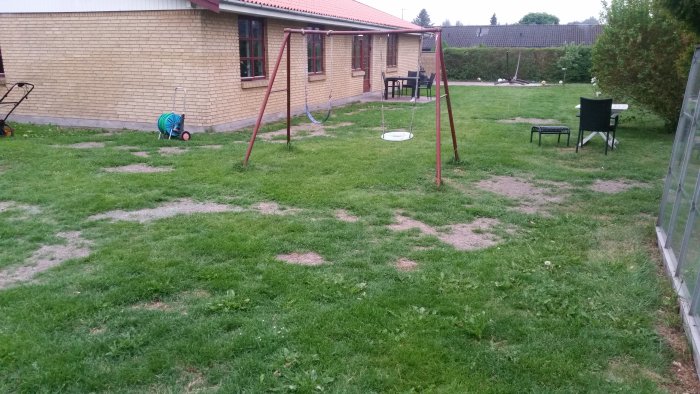 En trädgård med ojämn gräsmatta och fläckar av bar jord, gungställning och utemöbler i bakgrunden.