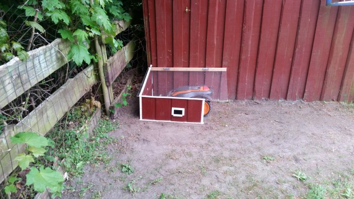 Robotgräsklippare under ett hemmagjort skyddstak vid en rödfärgad trävägg.