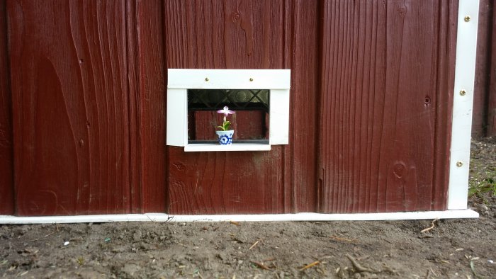 Hemgjort litet robotgräsklipparhus med ett fönster och en blomkruka.