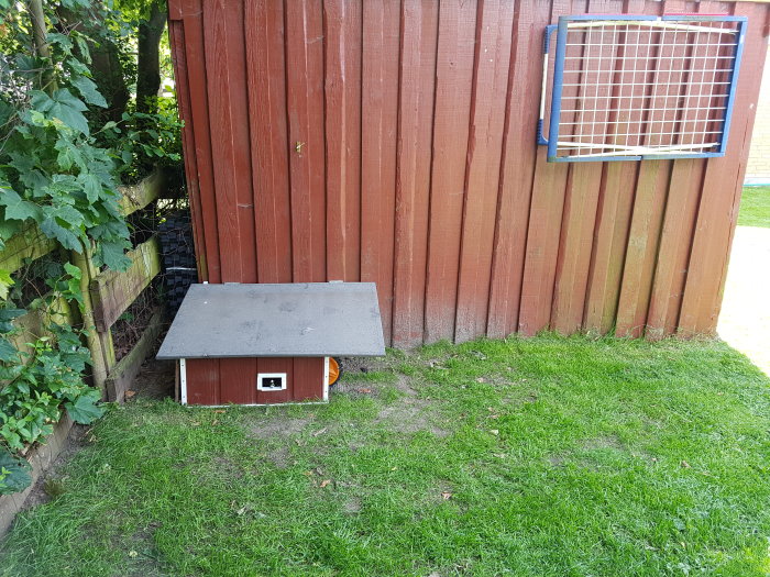 Ett litet trädgårdsprojekt med hemmagjord skyddshus för robotgräsklippare vid ett rött staket.