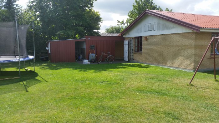 Trädgård med välskött gräsmatta, trampolin, cykel, och förrådshus bredvid ett tegelhus.