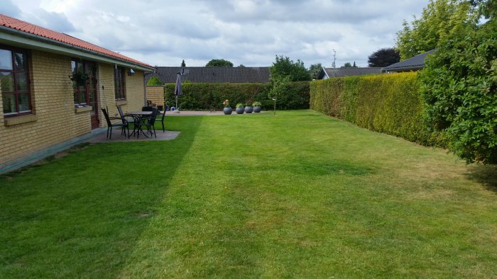 Välunderhållen gräsmatta bredvid en gul tegelvilla, med uteplatsmöbler och prydnadskrukor nära häcken.