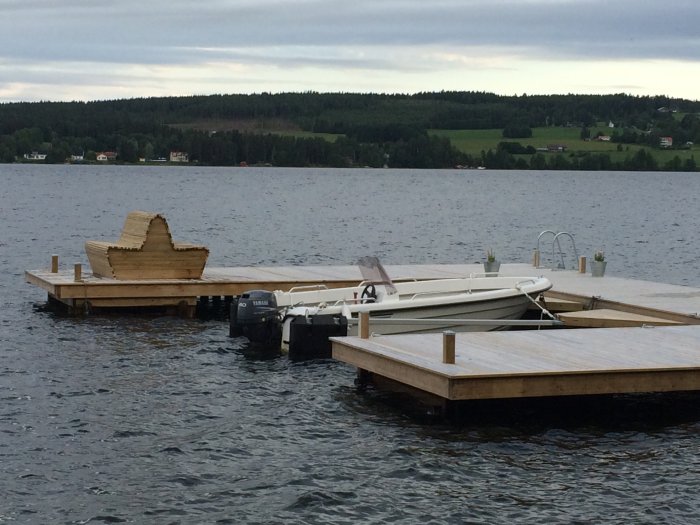 Handbyggd träsoffa på en brygga vid en sjö med båtar och lantlig bakgrund.