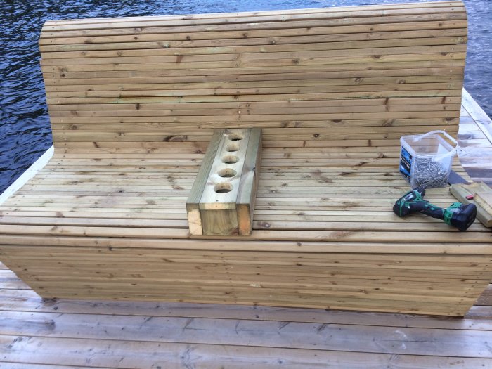 Träbänk vid vattnet med inbyggda dryckeshållare och verktyg utspridda på sidan.