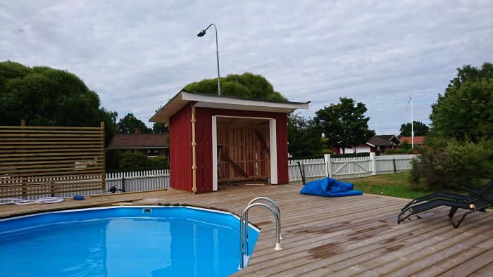 Nybyggt rött trädgårdsskjul med pappat tak och vita foder vid öppningen, bredvid en blå pool.