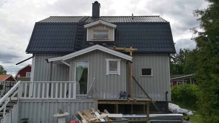 Grått hus under renovering med nytt fönsterfoder och uppställda trappstegar, samt material på en träveranda.