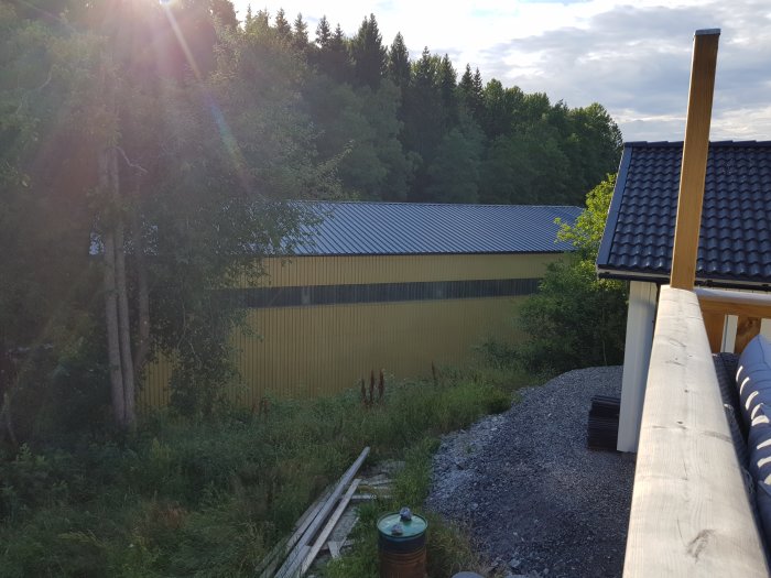 Vy över gul plåtbyggnad i släntläge sedd från en bostad, med träd och solfläckar.