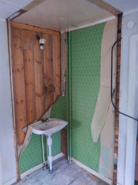 Hörn av äldre hus med delvis riven vägg, synlig timmerstomme och handfat med grön retro tapet.