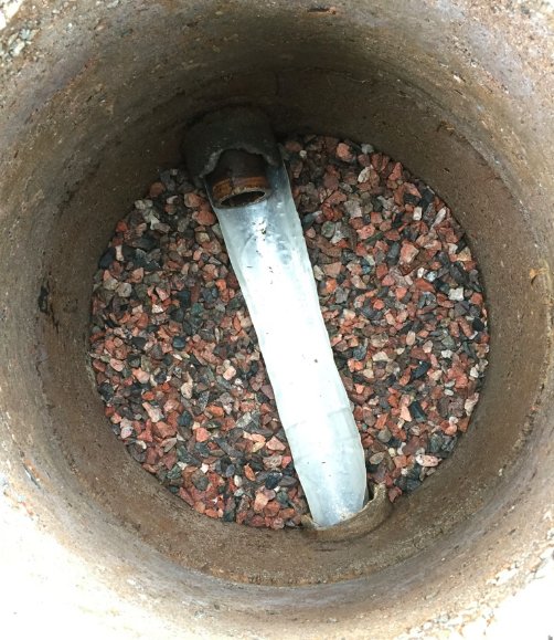 Äldre tvåkammarbrunn fylld med makadam och en provisorisk aluminiumplåtränna, väntar på anslutning till avlopp.