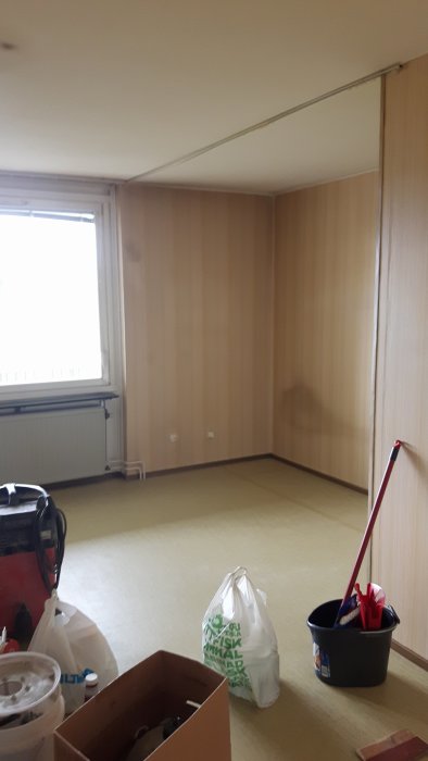 Tomt utdaterat rum med panelväggar inför renovering, med packade väskor och städmaterial på golvet.