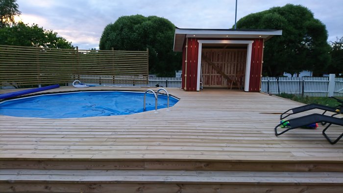 Utomhusvy över en trädäck med pool och rödmålat trädgårdsskjul vid skymningen.