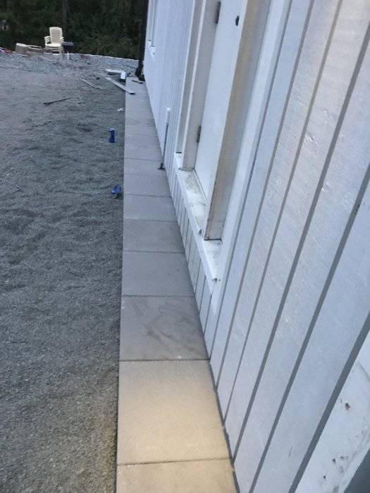 Första raden av stenplattor lagd längs husvägg med grusunderlag, förberedelse för montering av markrännor.