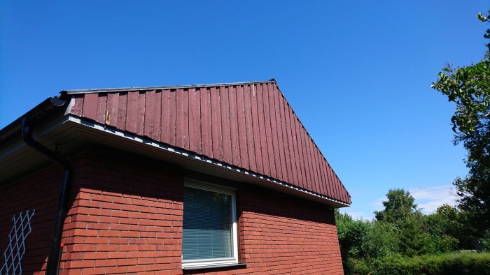 Hörnet av ett rött tegelhus med sliten träläkt panel på vinden under ett svart plåttak mot en klarblå himmel.