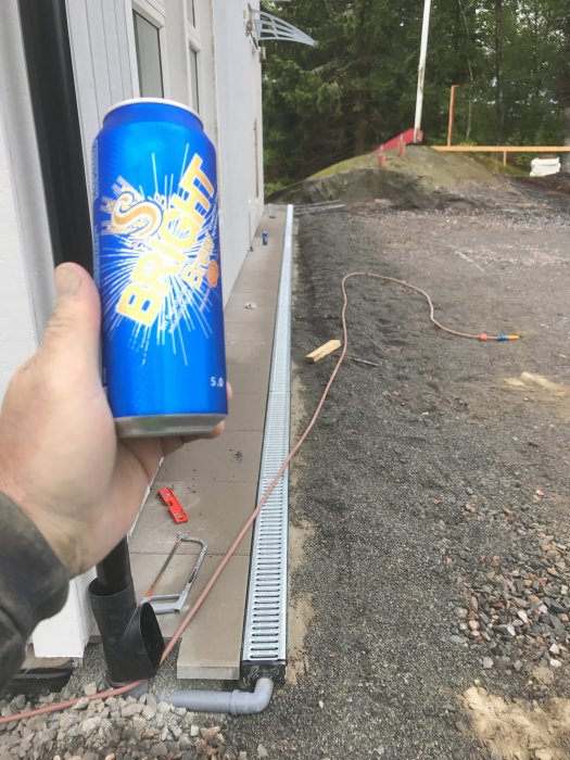 Hand håller dryckesburk framför nyinstallerad markränna vid husgrund med byggmaterial och jord i bakgrunden.