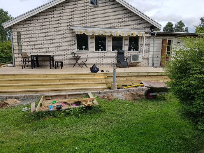 Nyuppförd trästolpe vid trappa på ett pågående däckbygge vid hus med trädgård, skottkärra och leksaker.