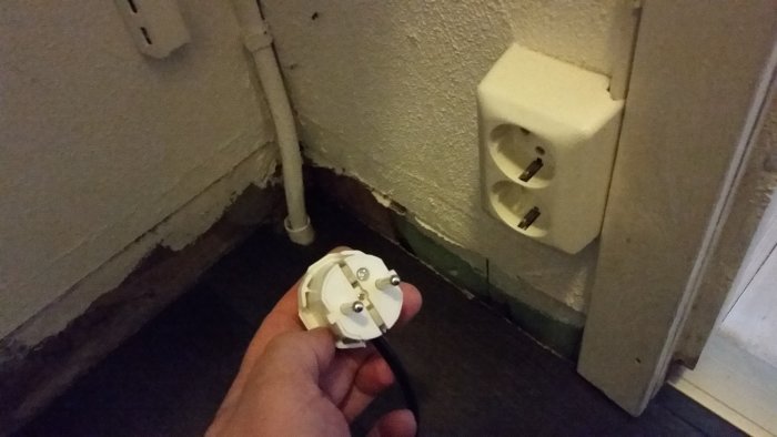 Hand håller stickpropp framför en oanvänd väggkontakt för provisorisk golvvärmeanordning.