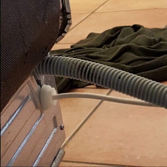 Vattenslang ansluten till vägguttag, nära golvet i ett kök, med oordnad grön trasa i bakgrunden.