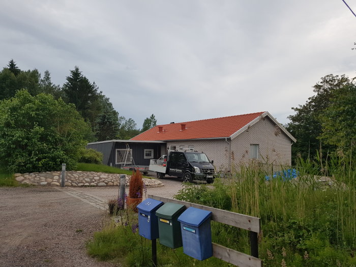 Husfasad med groventré, garage och parkerad firmabil, brevlådor i förgrunden och grönska.