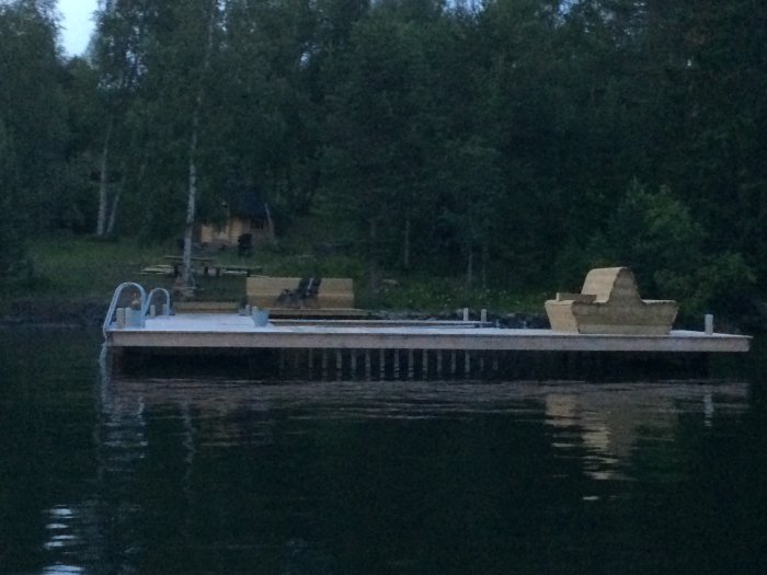 Morgonvy från sjön mot en brygga med bänkar och trappa, skymning, med skog och stuga i bakgrunden.