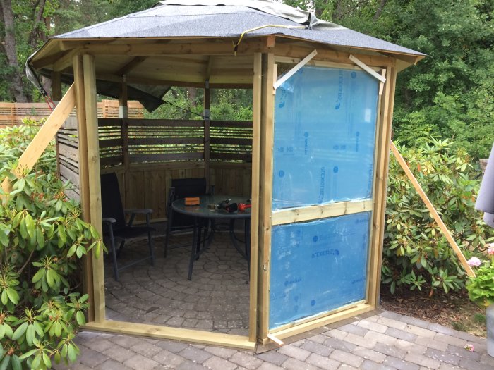 Byggprojekt av ett fönster med träram och plexiglas inuti en halvfärdig trädgårdsstuga på stenlagd mark.