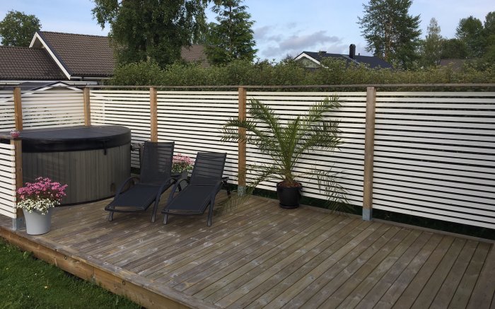 Trädäck med badtunna, solstolar och en sommarpalm till höger, intill vit staketvägg.