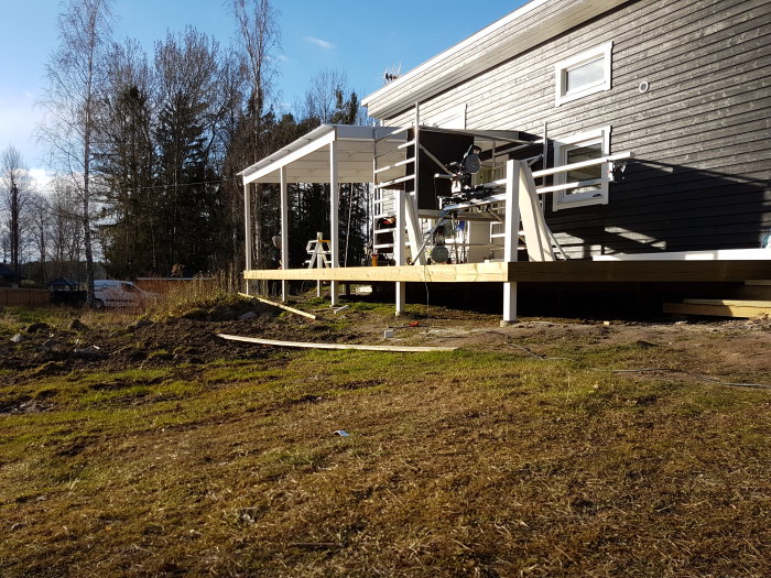 Pågående bygge av träaltan vid ett modernt hus, med verktyg synliga och delvis snötäckt mark.