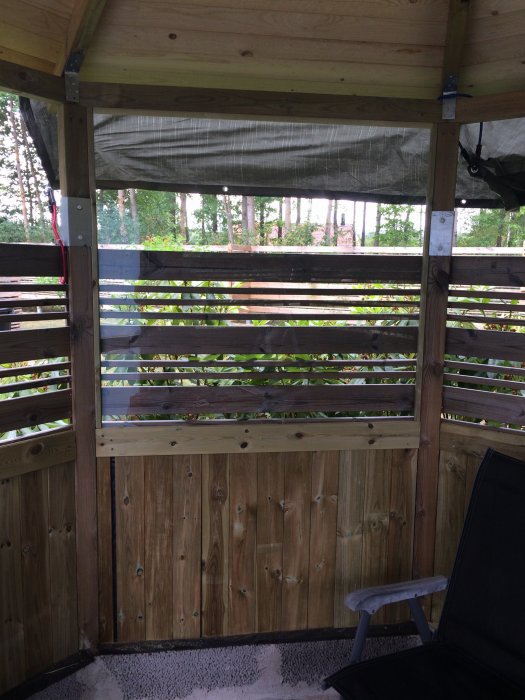 Träveranda med en installerad, vertikal vindruta ovanför en träpanelvägg, omsluten av trästaket och tak.