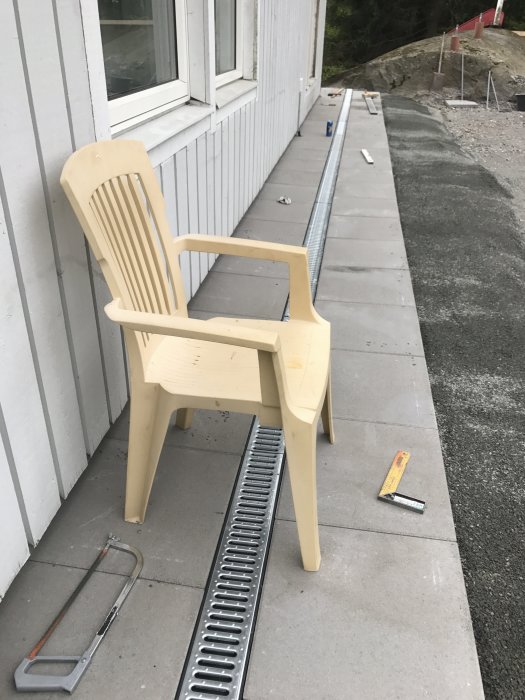 En beige vilostol står framför ett hus i byggfas, med påbörjad plattsättning och verktyg på marken.