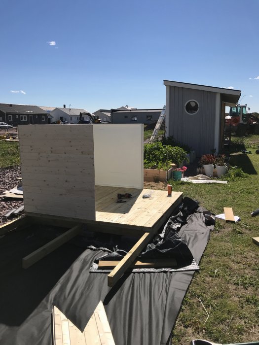 Halvfärdig lekstuga under konstruktion med nybyggt trädäck omgiven av en gräsplätt och byggmaterial.