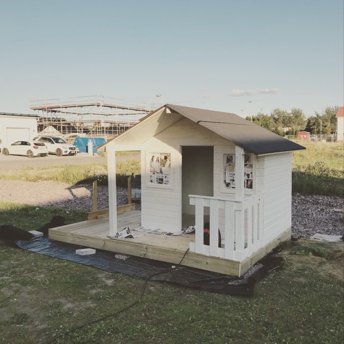 Nybyggd vit lekstuga med ett litet trädäck på en grusig tomt, med byggmaterial synligt.