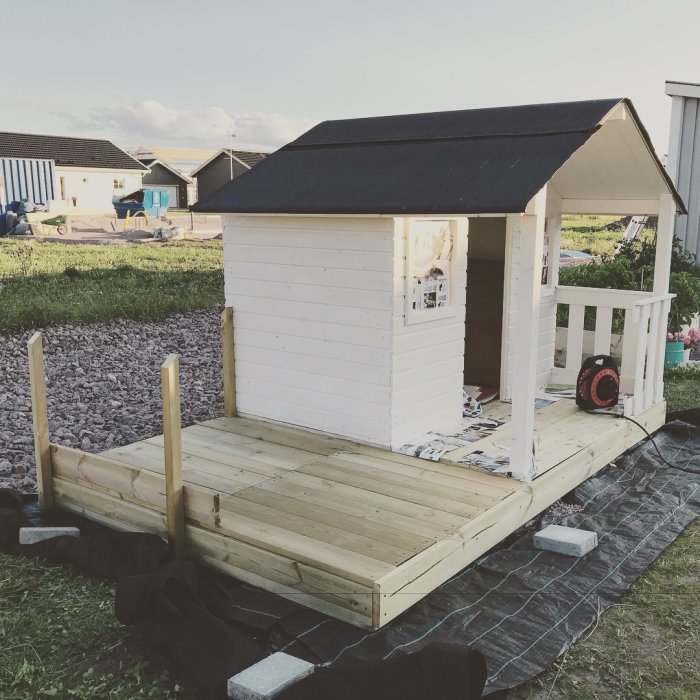 Nästan färdigbyggd lekstuga med tillbyggt trädäck i en trädgård, med verktyg synligt.