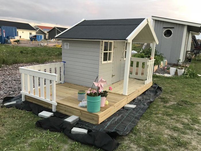 Nybyggd lekstuga med grå fasad och vitt trädäck, under uppbyggnad med skyddsduk och stenplattor runtom.