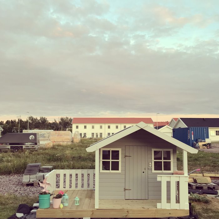 Nyligen byggd lekstuga med trädäck framför och rosa inredningsdetaljer, i början av skymningen.