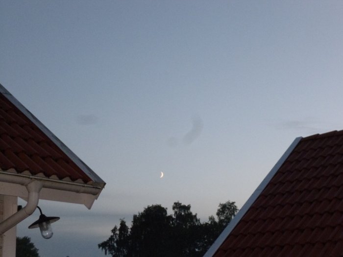 Kvällshimmel med nymåne mellan två hustak och subtila molnstråk som föreställer älvdans.