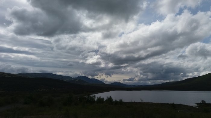 Vy över en sjö med norska fjällen i bakgrunden och moln på himlen, lite snö syns på fjälltopparna.