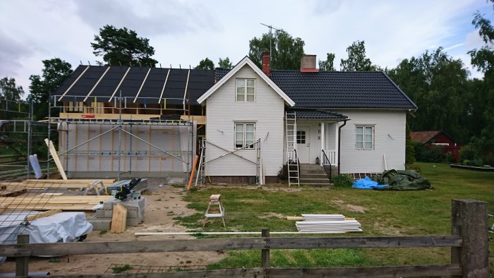 Hus under tillbyggnad med nya takstolar på ena sidan, byggmaterial framför och blå himmel i bakgrunden.