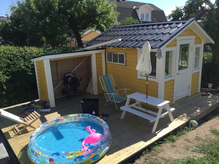 Gul lekstuga under renovering med öppet förrådsutrymme, omgiven av gräsmatta och en liten pool framför.