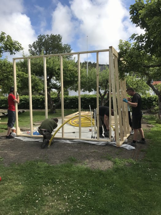 Personer reser trästommen till en byggnad i en trädgård på en solig dag.