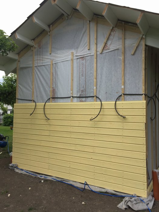 Delvis färdig fasad med gult grundmålat trä och vindskyddsduk på ett hus under uppbyggnad.