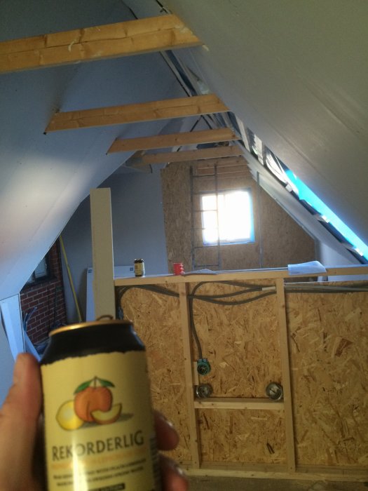 Renovering pågår i ett rum med oslipade träbjälkar och OSB-skivor, förgrund visar en hand som håller en cider.