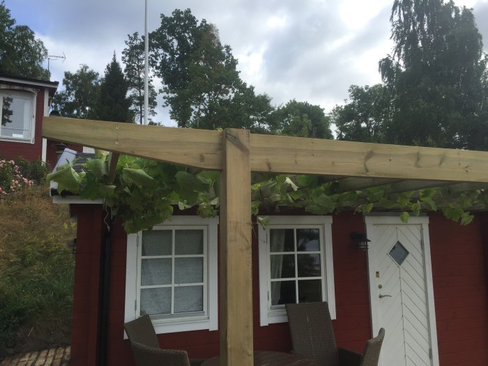 Renoverad altan med pergola och återbrukade träreglar, inbäddad i grönska intill ett rött hus.