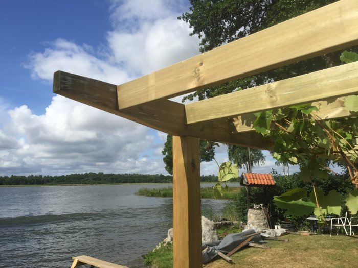 Nybyggd pergola med återanvänt trä och klättrande vinrankor, vid en sjökant med utsikt över vatten och träd.