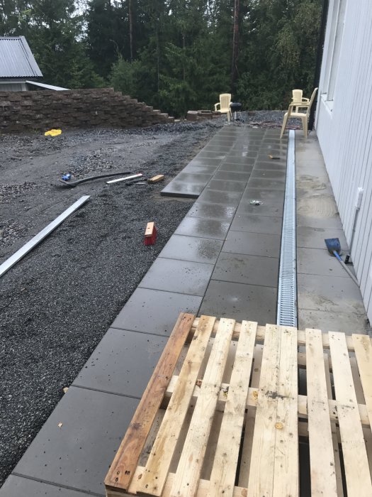 Pågående markarbete med stora, grå plattor och byggmaterial längs en husvägg.